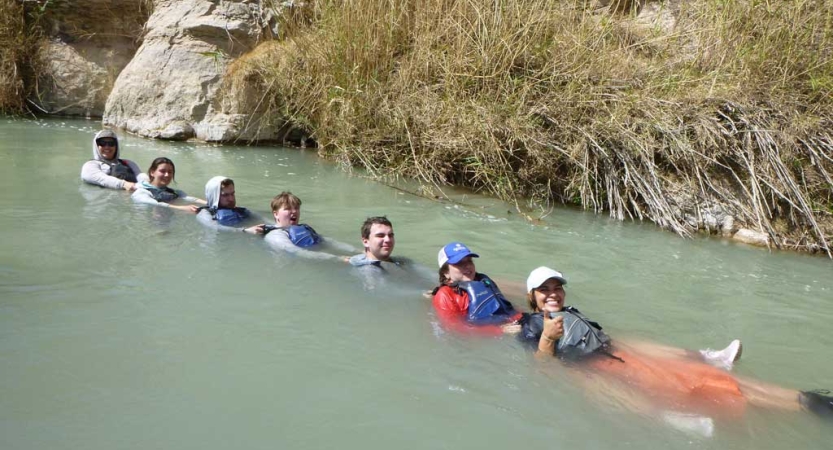 A group of people wearing life jackets float in water and hold onto each others' shoulders. 